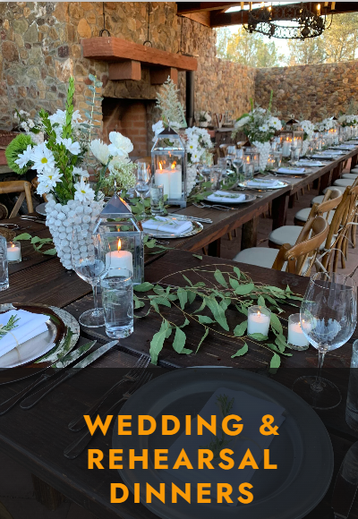 Elegant wedding and rehearsal dinner setup with white flowers, candles, and glassware on rustic wooden tables outdoors.